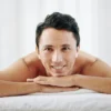 A man lays on his front on a treatment bed at a beauty salon, he rests his chin on his hands and smiles