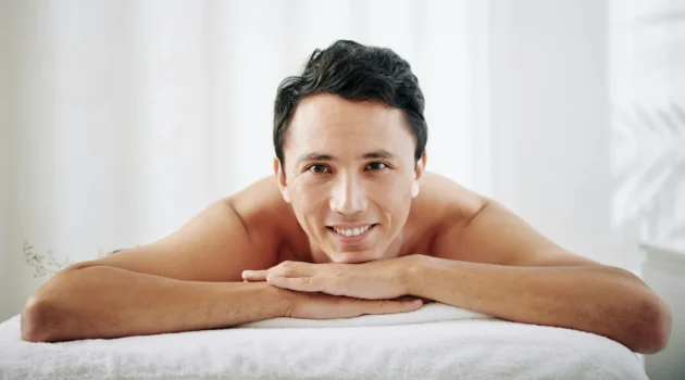 A man lays on his front on a treatment bed at a beauty salon, he rests his chin on his hands and smiles