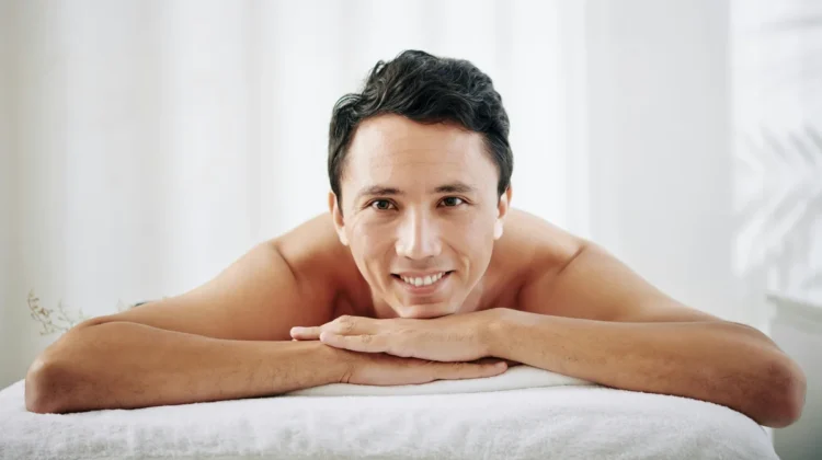 A man lays on his front on a treatment bed at a beauty salon, he rests his chin on his hands and smiles