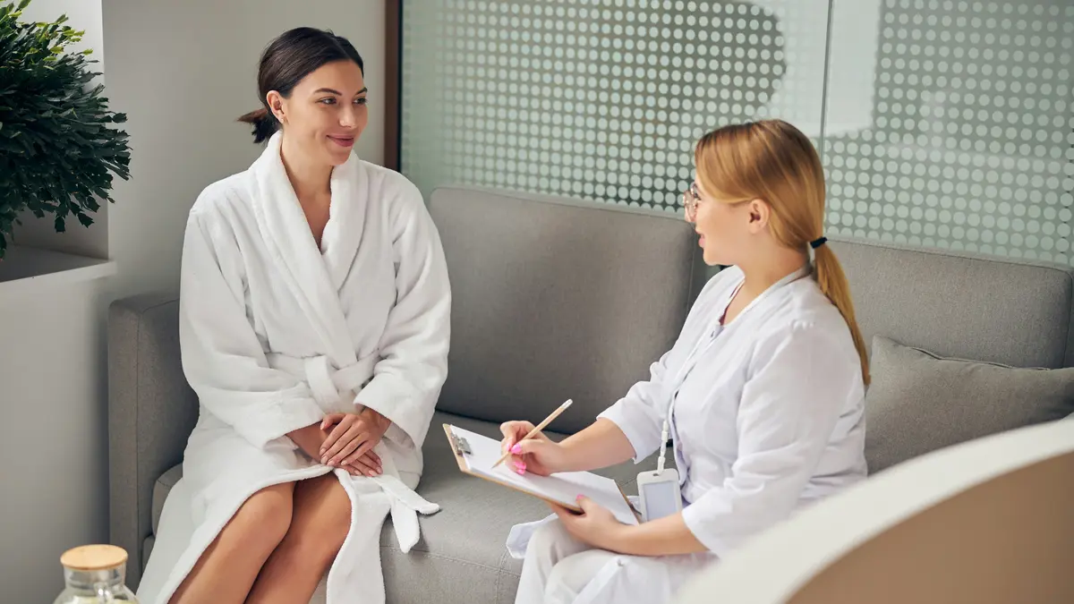 A client wearing a white robe and a therapist holding a clipboard sit next to each other on a sofa in a beauty salon as the therapist asks pre-treatment questions and writes them down